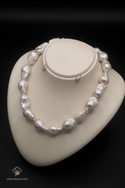 گردنبند مروارید باروک سفید با دانه‌های درشت و نامتقارن ۱۲ تا ۲۰ میلی‌متری روی استند کرم‌رنگ، پس‌زمینه مشکی